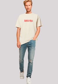 Cream-colored oversized t-shirt with red pixelated "SAIRU" text, paired with light blue distressed jeans and white sneakers.