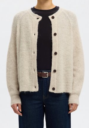 Personne portant un cardigan en maille beige avec des boutons foncés sur une chemise noire, un jean bleu et une ceinture marron avec une boucle argentée.