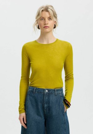 Young woman with blonde hair wearing a mustard yellow long-sleeve top and high-waisted blue jeans, standing against a plain background.