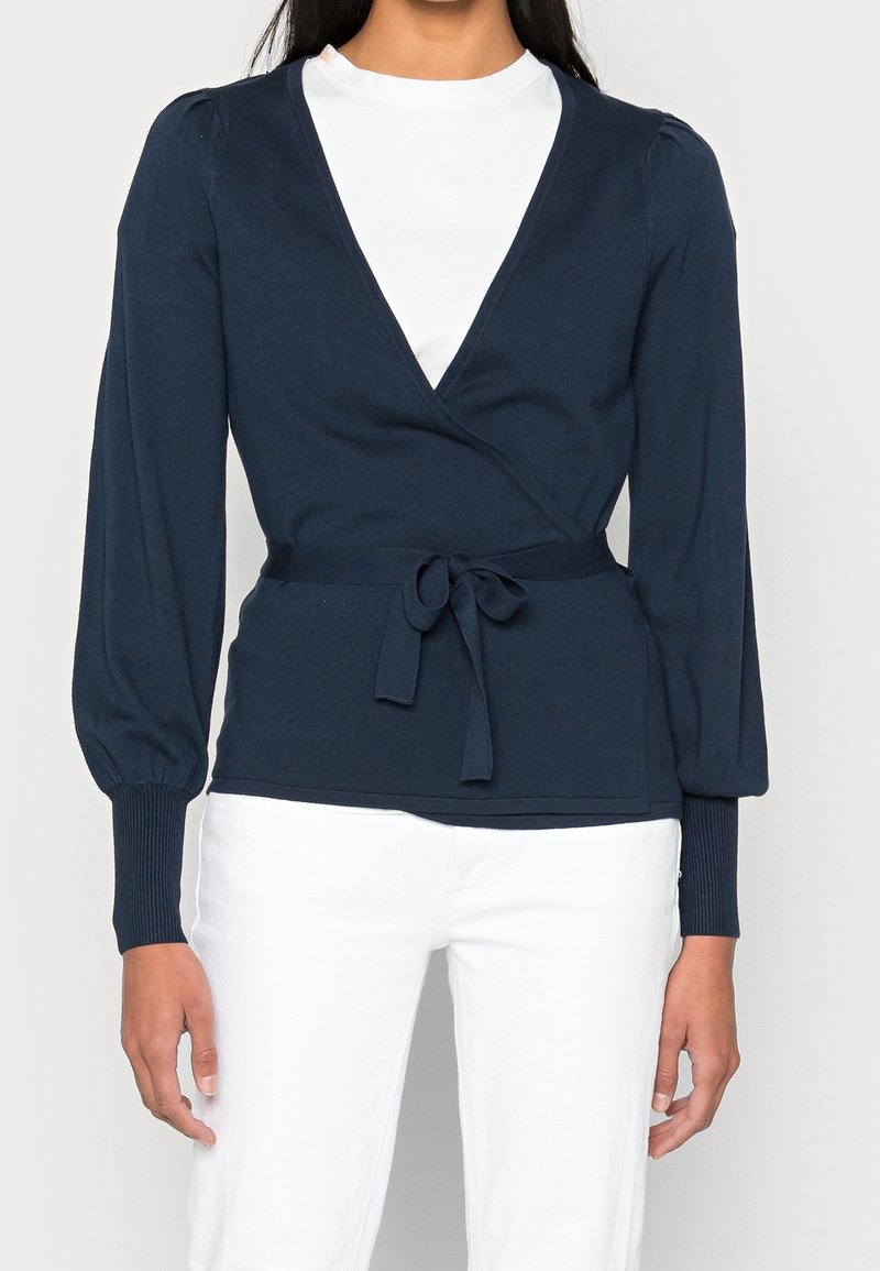 Femme portant un pull cache-cœur bleu marine avec une ceinture nouée sur un top blanc et un pantalon blanc, vue du cou aux hanches.