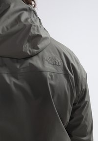 Personne portant une veste imperméable vert olive avec une capuche, affichant le logo "The North Face" brodé sur le haut du dos.