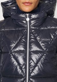 Mörkblå, glansig pufferjacka med huva, utrustad med dragkedja fram och mönster av diamantformad quiltning. Silverfärgade detaljer.