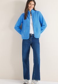 Femme portant une veste en polaire bleu vif, une chemise blanche, un jean bleu foncé et des baskets argentées, debout contre un mur de couleur claire.