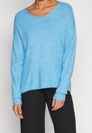 Personne portant un pull en maille bleu clair à manches longues avec un large col et un pantalon noir, debout devant un fond uni.