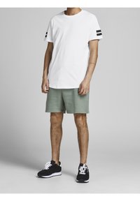 Camiseta blanca de algodón con rayas negras en las mangas, combinada con pantalones cortos verdes y zapatillas negras con suelas y cordones blancos.