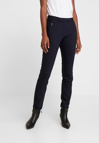 Pantalons bleus marine ajustés en tissu extensible, avec une finition lisse, une coupe slim et deux poches avant zippées. Portés avec des bottines noires.