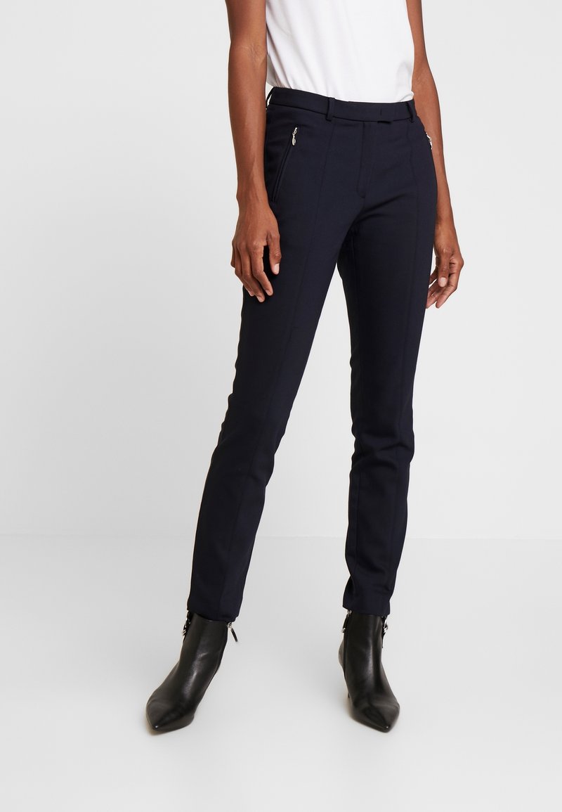 Pantalons bleus marine ajustés en tissu extensible, avec une finition lisse, une coupe slim et deux poches avant zippées. Portés avec des bottines noires.