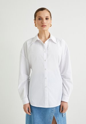 Hvid skjorte med knaplukning og klassisk krave, lange ærmer og løs pasform. Stoffet har en glat tekstur, der komplementeres af et denimskørt.