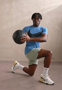 Sportists zilā Nike T-kreklā ar melnu logo, zaļos šortos un baltajās kurpēs, turot melnu medicīnas bumbu, ceļos uz pelēka grīdas.