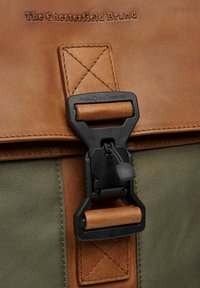 Rabats en cuir brun avec texte en relief et boucle magnétique Fidlock noire. Présente un tissu texturé vert en dessous, avec des coutures accentuées.