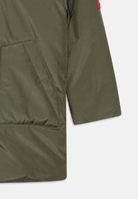 Chaqueta acolchada de color verde oliva con un bolsillo lateral, textura acolchada y corte recto. Presenta un detalle en rojo en la manga.