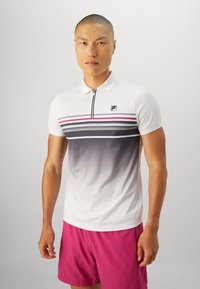 Man wearing a white short-sleeve polo with black and pink stripes and fuchsia shorts, standing against a plain light background.