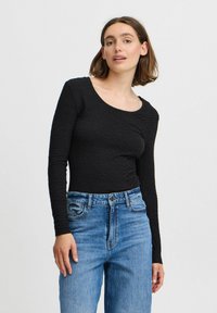 Črna dolgosleeve majica z teksturiranim vzorcem, okroglo izrezano ovratnikom, prilegne silhueto, v kombinaciji z visokim pasom modrimi kavbojkami.