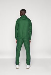 Grønn treningsoverdel med hette, elastiske mansjetter, matchende bukser med blå sidestreker, og hvite joggesko. Glatt tekstur, sporty design.