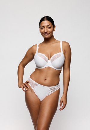 Vrouw met donker haar in een witte kanten beha en bijpassende slipjes staat zelfverzekerd tegen een effen witte achtergrond.