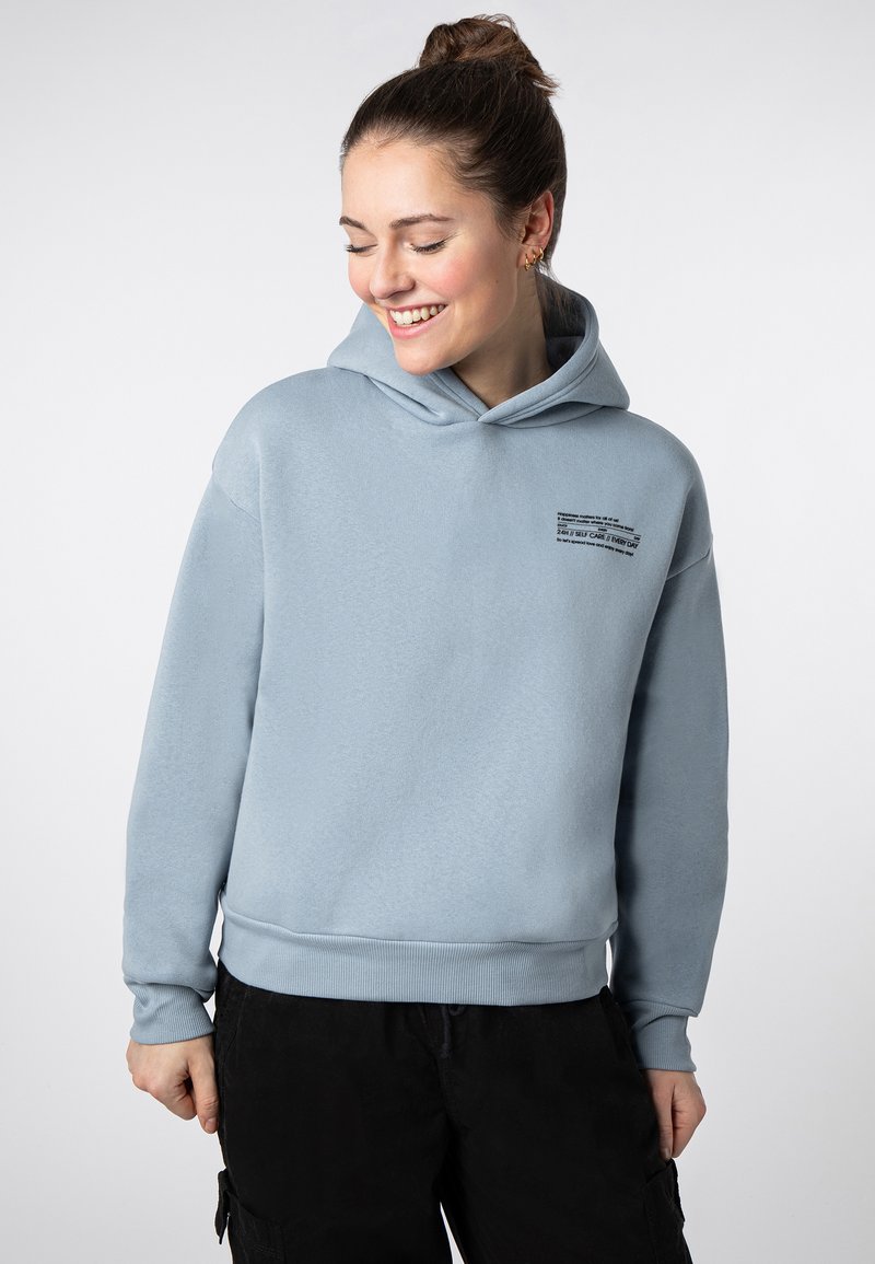 Hellblauer Kapuzenpullover mit lockerem Schnitt, der eine kleine schwarze Textdruck-Detail auf der Brust sowie gerippte Bündchen und Saum aufweist.