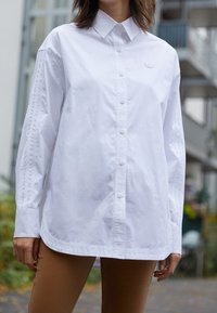 Vit långärmad skjorta i bomull, med knäppning framtill, klassisk krage och diskret logotyp på bröstet. Slät textur.