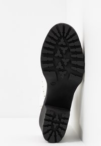Semelle d'une botte noire à semelle crantée avec des rainures profondes et des motifs triangulaires, positionnée verticalement sur un fond blanc.