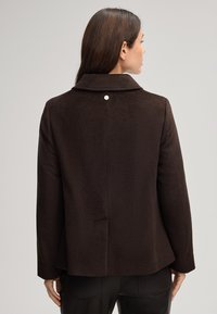 Cappotto in lana marrone con colletto, dettaglio di un singolo bottone sul retro e vestibilità rilassata. La texture liscia e la forma strutturata ne esaltano il design.