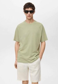 Zelena majica s kratkim rukavima od mekanog materijala, u kombinaciji sa svijetlo bež lanenim šortsevima. Model nosi crne sunčane naočale, ruke u džepovima.