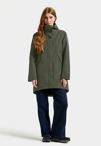 Olivgrön vattentät jacka med hög krage, dragkedja framtill och två sidfickor, kombinerad med mörkblå vida jeans och svart skor.