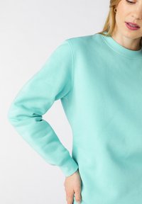 Felpa turchese chiaro con polsini a costine e collo rotondo. Texture liscia, cuciture visibili e silhouette rilassata.