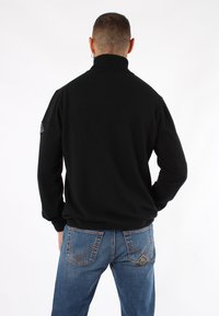 Maglione a collo alto nero e a coste realizzato in un tessuto morbido, abbinato a jeans blu con modelli di cuciture distintivi e tasche.