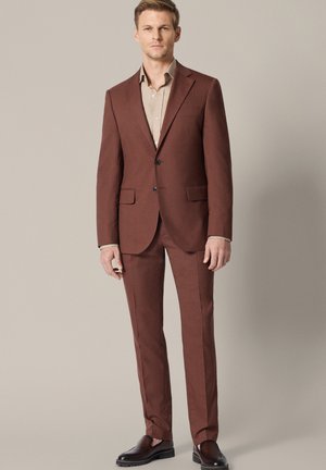 Mężczyzna stoi ubrany w dopasowany brązowy garnitur z beżową koszulą i ciemnobrązowymi eleganckimi butami na jednolitym beżowym tle.