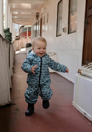 Väikelaps kalamustris sinises kombinesoonis ja mustade saapastega jalutab ja naeratab laeva koridoris, kus on valged seinad ja puituks.