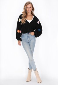 Svart stickad cardigan med lösa ärmar, prydd med färgglada blomapplikationer, i kombination med ljusblå slitna jeans och beige ankelskor.