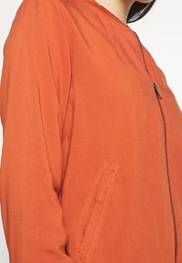 Oranžová bomber bunda s ribbeným límcem, bočním kapsou a plným zipem. Hladká textura, lehký materiál.