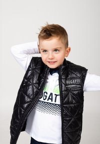 Gilet trapuntato nero con una texture lucida, indossato su una camicia bianca con disegni verdi e blu navy, e jeans blu. Logo visibile sul gilet.