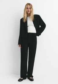 Kvinna med långt blont hår klädd i en svart oversized kavaj, vit kort topp, svarta vida byxor och svarta platta skor mot en vit vägg.