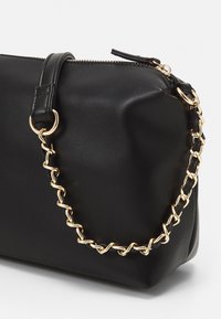 Borsa a tracolla in pelle nera con dettaglio della catena dorata e chiusura con cerniera superiore su sfondo bianco.