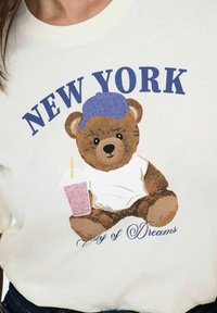 Cremefarvet t-shirt med en tegneserie-bjørn i blå hat, der holder en lyserød drink. Teksten siger "NEW YORK" og "Byen med Drømme" nedenunder.