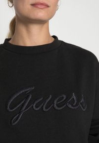 Vrouw in een zwarte crewneck sweatshirt met een verhoogd geborduurd "Guess"-logo op de borst en een gouden oorring zichtbaar aan het linkeroor.