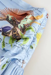 Tissu bleu clair avec un imprimé girafe coloré et plantes tropicales, comprenant un texte en cursive noir et un détail de ceinture élastiquée froncée.