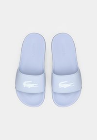 Sandales slide en caoutchouc bleu clair avec une large sangle ornée d'un logo crocodile blanc en relief. Surface lisse, bout arrondi.