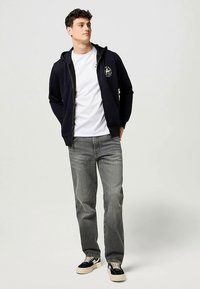 Sweat à capuche zippé bleu marine avec poches à l'avant, détail du logo sur la poitrine, associé à un t-shirt blanc et un jean gris, porté avec des baskets noires.