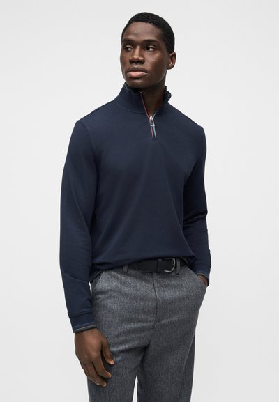 Μπλε σκούρο πουλόβερ με μακριά μανίκια και φερμουάρ στον λαιμό, με αντίθετη λεπτομέρεια στο γιακά, συνδυασμένο με γκρι υφασμένα παντελόνια και μαύρη ζώνη.