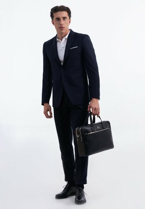 Completo blu con una polo bianca, valigetta in pelle nera e scarpe eleganti nere. La valigetta ha una cerniera e dettagli hardware in argento.
