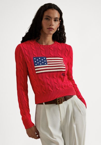 Pulover roșu cu model tricotat în formă de cablu, având un steag american brodat pe partea din față și manșete și tiv drepte. Asortat cu pantaloni crem cu talie înaltă.