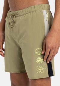 Bañadores de hombre en color verde oliva con una cintura elástica y cordón. Presentan gráficos amarillos y bolsillos laterales con acentos negros contrastantes.