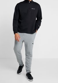 Pile nero con mezza cerniera e logo bianco, abbinato a pantaloni della tuta in cotone grigi con tasca laterale e dettagli neri.