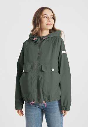 Vrouw draagt een donkergroene ritssluiting hoodiejack met klepzakken aan de voorkant en roze trekkoorden, gecombineerd met een blauwe spijkerbroek.