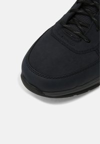 Black athletic shoe with a smooth, matte leather upper and black mesh detailing. Features a textured rubber sole and black lace closures.