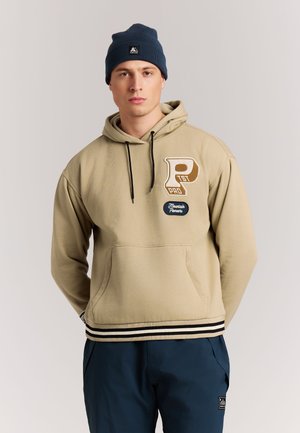 Man wearing beige hoodie with patches and black drawstrings, navy blue pants, and navy beanie standing against light background.