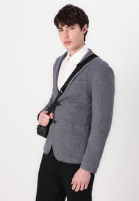 Γκρίζο ριγέ σακάκι με δύο μπροστινές τσέπες, κλείσιμο με έναν μόνο κουμπί και υφή, συνδυασμένο με λευκή πουκαμίσα με γιακά και μαύρη τσάντα χιαστί.