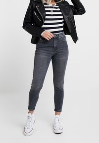 Kobieta w czarnej skórzanej kurtce, czarno-białej w paski koszulce, ciemnoszarych jeansach skinny z wysokim stanem oraz białych trampkach.