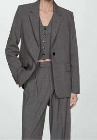 Frau trägt einen grauen Nadelstreifen-Dreiteiler mit hoch taillierter, weit geschnittener Hose, geknöpfter Weste und übergroßem Blazer vor einfachem Hintergrund.